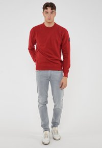 Roter, gestrickter Pullover mit Rundhalsausschnitt, getragen über hellgrauen Jeans. Verfügt über ein kleines Logo-Patch an der linken Schulter.