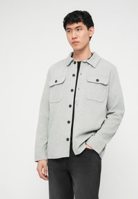 Νέος άνδρας που φοράει ανοιχτό γκρι σακάκι με κουμπιά πάνω από μαύρο πουκάμισο, με το ένα χέρι στην τσέπη σκούρου γκρι παντελονιού, στέκεται μπροστά σε λευκό φόντο.