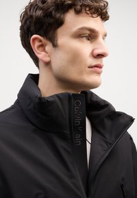 Giacca nera con zip e colletto alto, dotata di dettagli riflettenti lungo la cerniera e una texture liscia e opaca. Design minimalista.