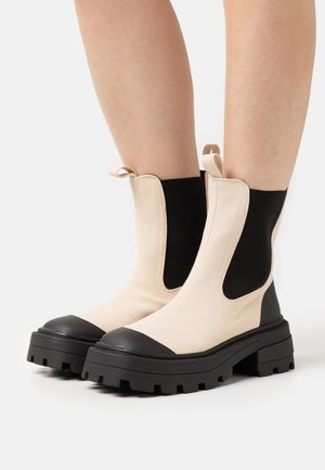 Botas de tornozelo chunky em bege e preto, feitas de material suave com painéis laterais elásticos e uma sola espessa e texturizada.
