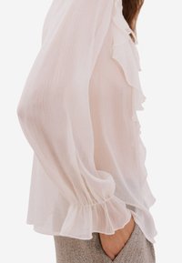 Světlá růžová halenka s volánky vpředu, průsvitná látka, nařasené manžety a volný střih, doplněná texturovanými béžovými kalhotami.