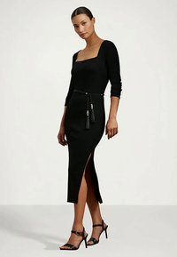 Frau in einem eng anliegenden schwarzen Midikleid mit Dreiviertelärmeln, quadratischem Ausschnitt, seitlichem Schlitz, Quasten-Gürtel und schwarzen High Heels.