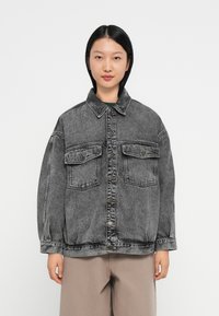 Mulher com cabelo preto liso, vestindo uma jaqueta de denim preta desbotada de corte solto, com botões à frente e bolsos de aba, combinada com calças bege.