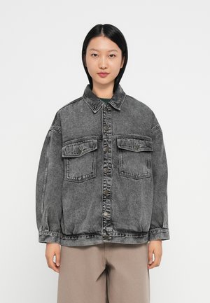 Kvinne med rett svart hår som har på seg en løs, falmet svart denimjakke med knappelukking og klaffelommer, sammen med beige bukser.