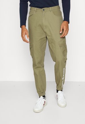 Mann trägt olivgrüne Cargo-Joggerhosen mit elastischen Bündchen, weiße Sneakers, schwarze Socken und ein langärmeliges dunkelblaues Shirt und steht auf einem weißen Boden.