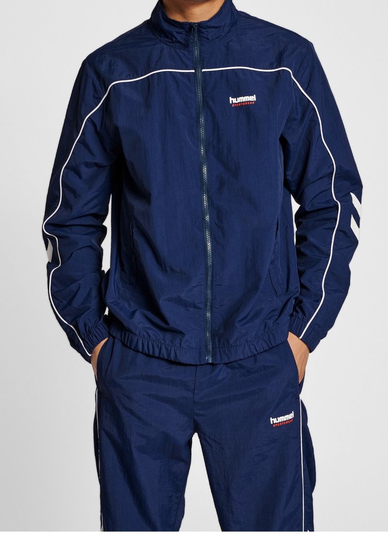 Man wearing a navy blue hummel sportswear tracksuit with white piping, hands in pockets, standing against a plain background.