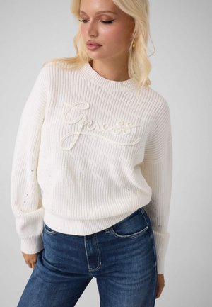 Femme blonde portant un pull en tricot blanc avec un logo en relief "Guess" et un jean bleu, regardant vers le bas et légèrement sur le côté sur fond uni.
