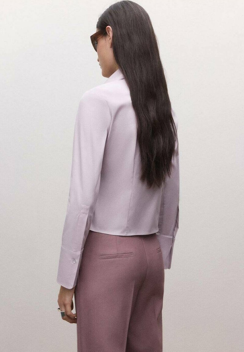 Femme aux longs cheveux foncés portant des lunettes de soleil, un chemisier rose clair et un pantalon mauve, debout devant un fond uni clair.