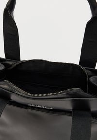 Μαύρη τσάντα tote με λεία, γυαλιστερή επιφάνεια και φερμουάρ στην κορυφή. Το εσωτερικό διαθέτει υφασμάτινη επένδυση και επιπλέον τσέπη με φερμουάρ.