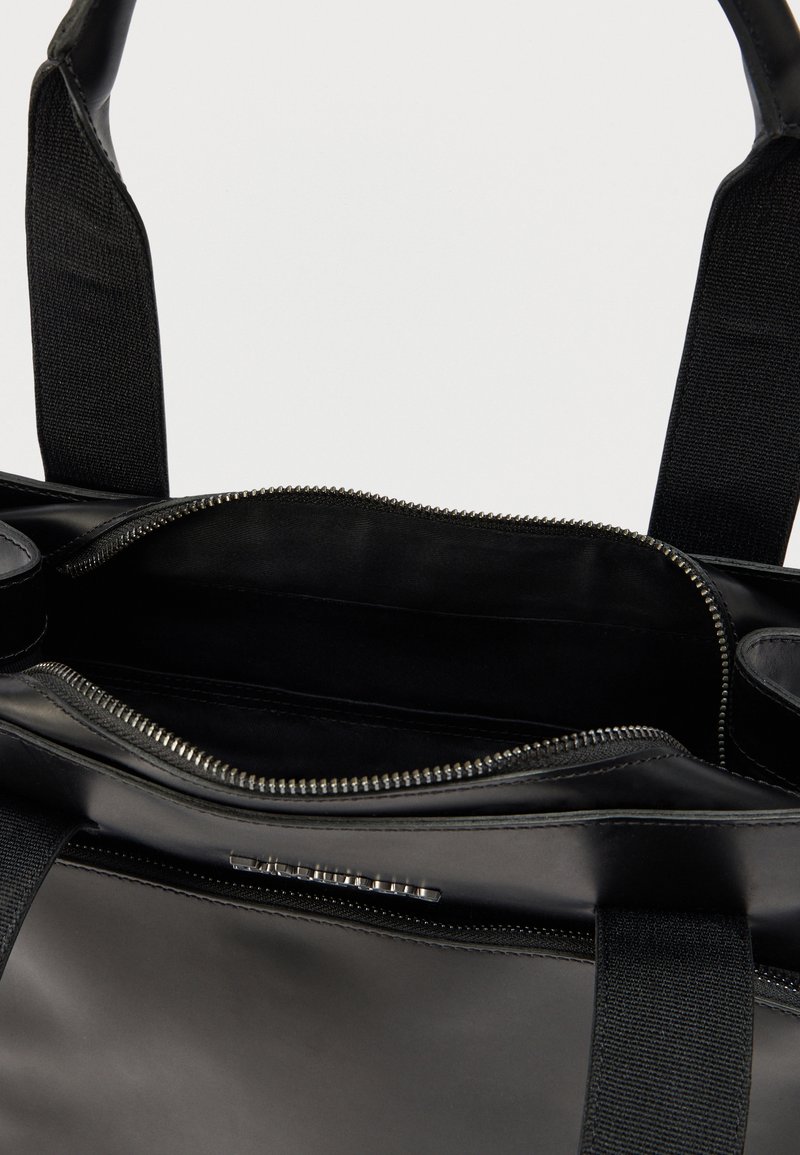 Zwarte draagtas met een glad, glanzend oppervlak en een ritssluiting aan de bovenkant. Het interieur is voorzien van een stoffen voering en een extra vak met ritssluiting.