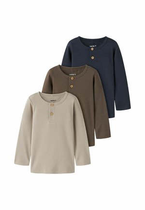 Trois chemises henley à manches longues en beige, marron et bleu marine, chacune avec deux boutons en bois au col rond, posées en chevauchement sur fond blanc.