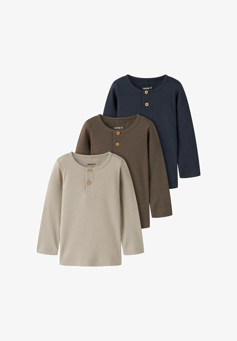 Trois chemises henley à manches longues en beige, marron et bleu marine, chacune avec deux boutons en bois au col rond, posées en chevauchement sur fond blanc.