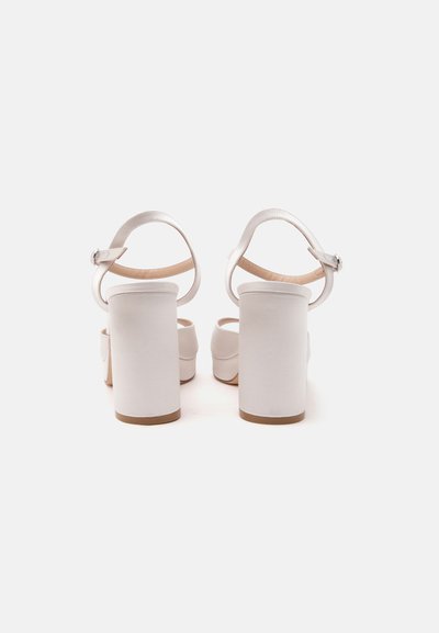 Paire de sandales blanches à talons hauts avec brides à la cheville et talons blocs vues de dos sur un fond clair uni.