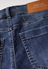 Jeans en denim dans un lavage indigo foncé, présentant des coutures contrastées, deux poches arrière et une étiquette en cuir marron avec un texte en relief.