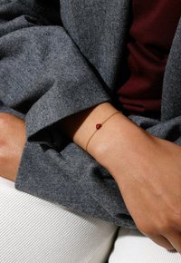 Gold chain bracelet featuring a red gemstone on a wrist, paired with a grey textured fabric and a light-colored ribbed surface below.