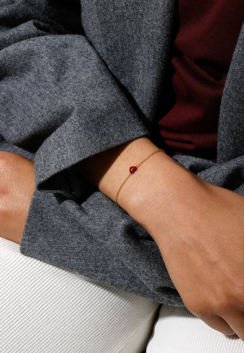 Gold chain bracelet featuring a red gemstone on a wrist, paired with a grey textured fabric and a light-colored ribbed surface below.