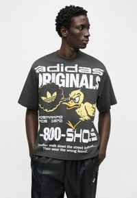 Άνδρας με μαύρο T-shirt Adidas Originals με μεγάλο λευκό και κίτρινο καρτούν πάπια γραφικό και μαύρο παντελόνι, στέκεται με τα χέρια στις τσέπες.