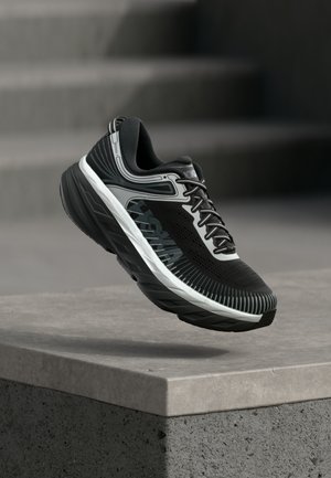 Čierna a sivá športová obuv s hrubou podrážkou, vznášajúca sa nad betónovým okrajom, s rozmazanými betónovými schodmi v pozadí.