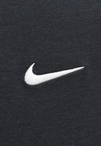 Juoda audinio medžiaga turi ryškų baltą išsiuvinėtą Nike Swoosh logotipą, demonstruojantį lygią tekstūrą ir vienodą mezgimo raštą.