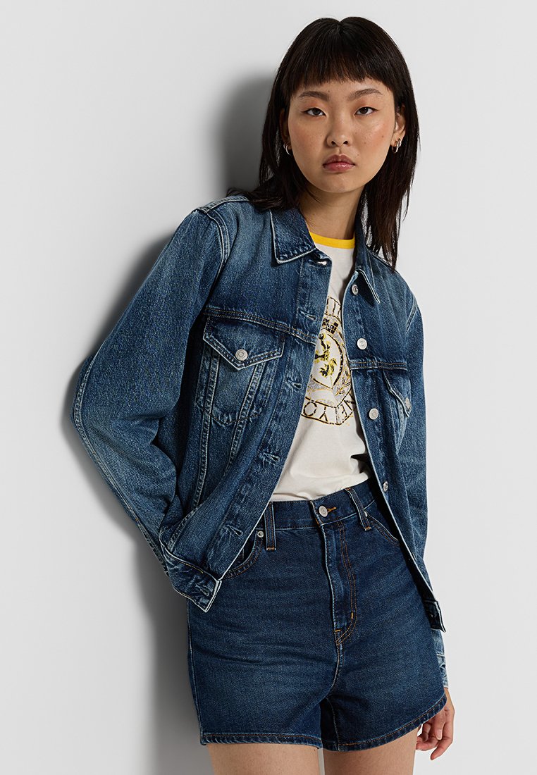 Dunkelblaue Jeansjacke mit klassischem Design, Knopfleiste, Brusttaschen und leichter Abnutzung. Kombiniert mit einem grafischen T-Shirt und dunklen Shorts.