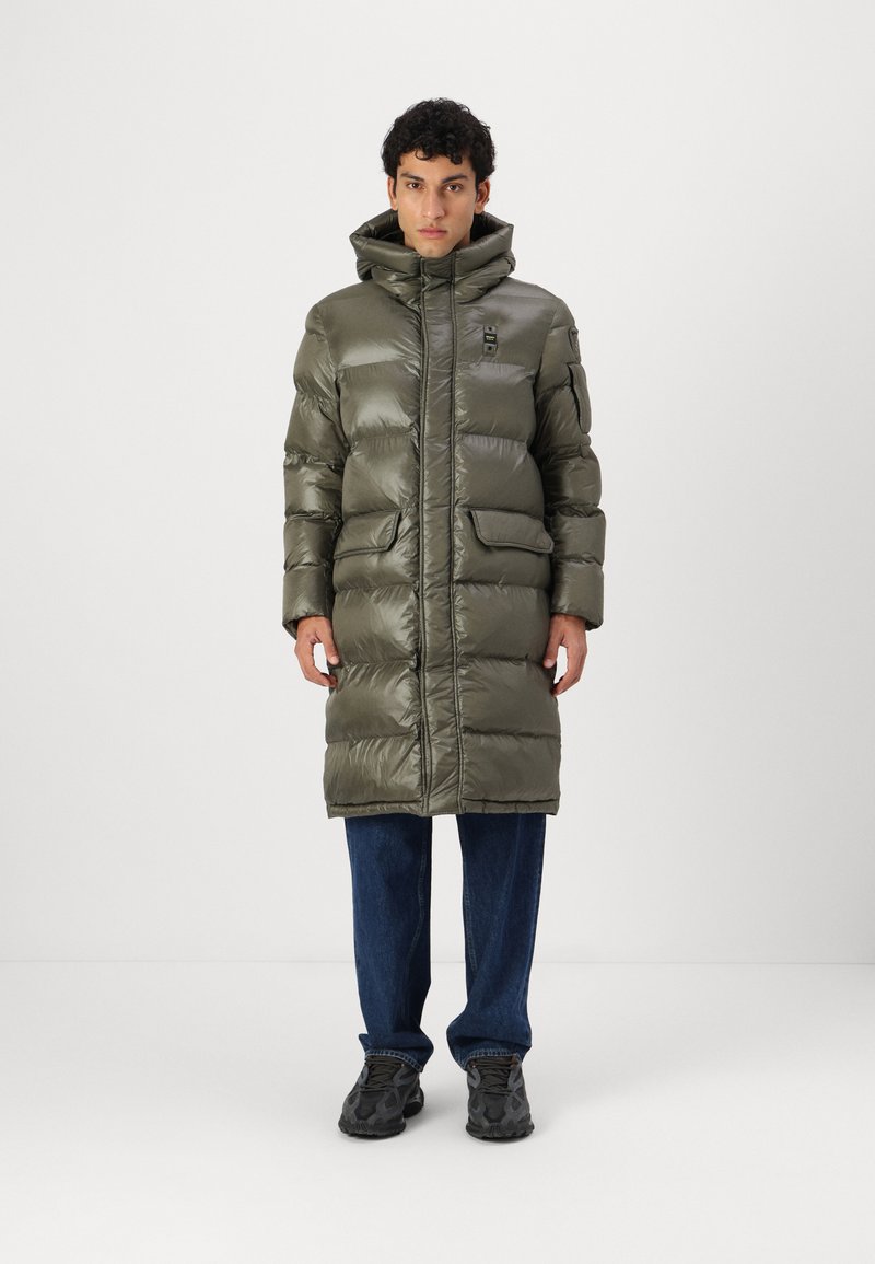 Puffy olive-green long coat with a high collar, front zip closure, and two side pockets. Worn over blue jeans and dark sneakers.