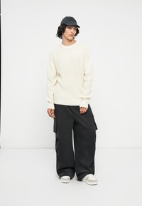Ljusgrå stickad tröja, lösa svarta cargobyxor med fickor, och en mörk keps. Vita sneakers med grå detaljer. Enkelt, avslappnat outfit.