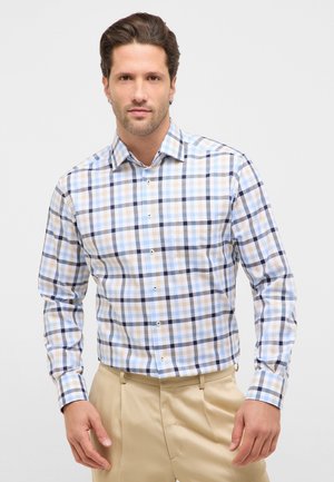 Mand iført en langtærmet skjorte med knapper i et mønster af blå, beige og sort tern og beige bukser mod en ensfarvet baggrund.