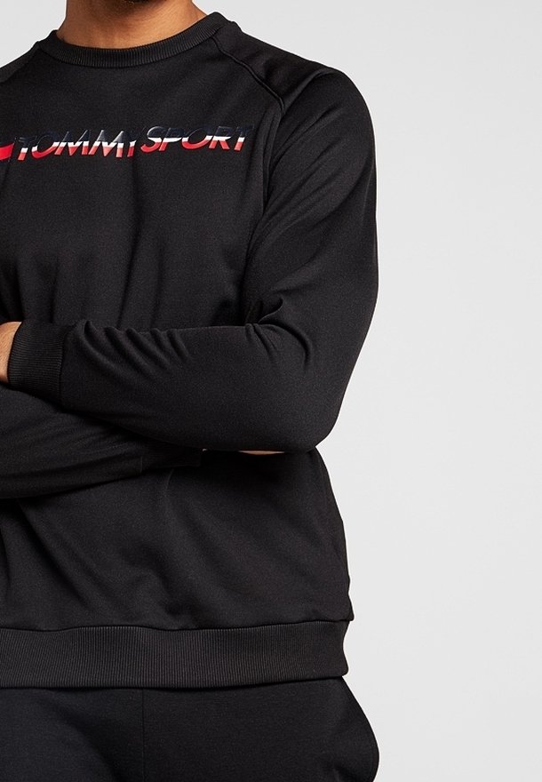 Personne portant un sweat-shirt noir à manches longues avec le logo "TOMMY SPORT" en rouge, blanc et bleu sur la poitrine, les bras croisés.