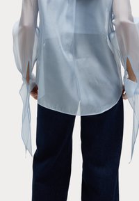 Blusa azzurro chiaro, semi-trasparente, con maniche larghe e fessure aperte; texture liscia; abbinata a pantaloni in denim scuri e larghi.