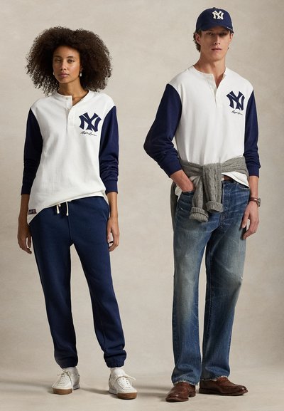 Un duo de mannequins portant des vêtements de sport décontractés. La femme porte une chemise à manches longues blanche et marine avec un logo et un jogging marine. L'homme porte une chemise similaire et un jean bleu, avec un pull gris noué autour de la taille.