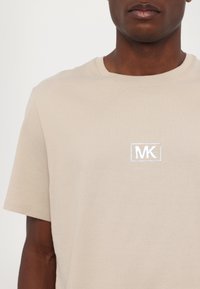 Lys beige t-shirt lavet af blød bomuld. Har korte ærmer, en rund halsudskæring og et hvidt broderet "MK" logo inden i en rektangulær ramme.