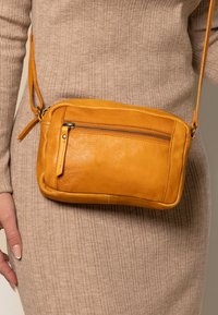 Kvinna som bär en ribbad beigemodell med en liten mustardgulfärgad lädercrossbody-väska med framficka med dragkedja.