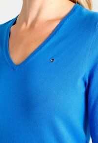 Sweter z dekoltem w serek, wykonany z jasno niebieskiego materiału, z małym logo marki umieszczonym na lewej piersi. Gładka tekstura, dopasowany krój.