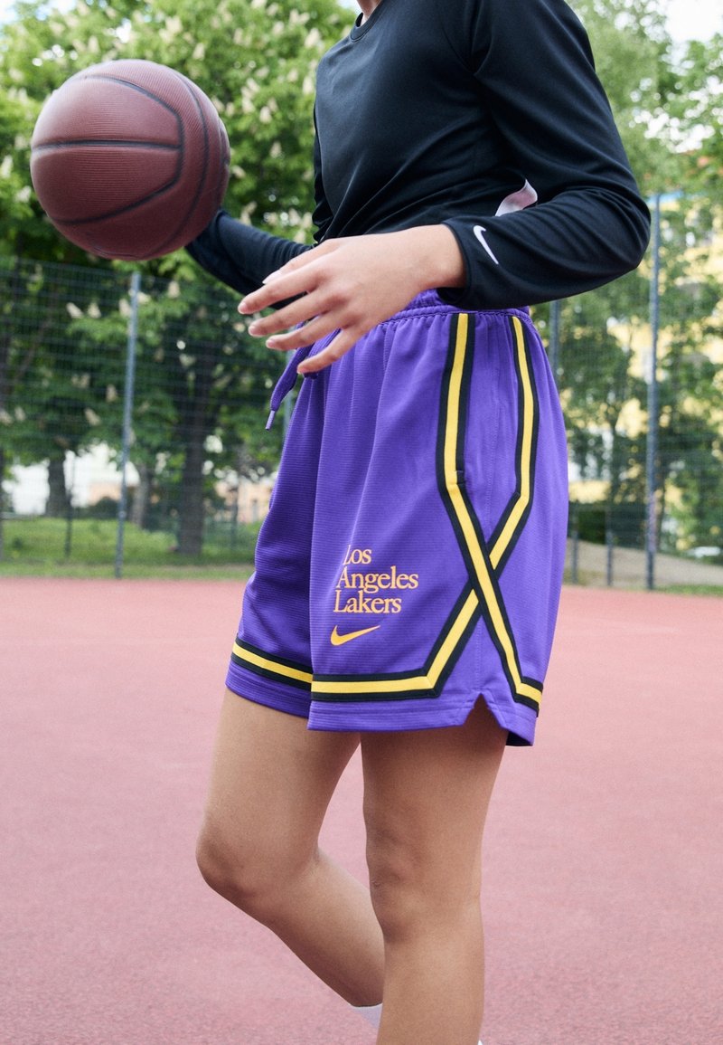 Personne dribblant un ballon de basket sur un terrain extérieur, portant un short violet des Los Angeles Lakers et un haut noir à manches longues.