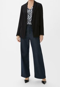 Blazer nero sopra una maglietta blu a motivi, abbinato a jeans scuri a gamba larga e décolleté neri a punta. Design semplice e sartoriale.