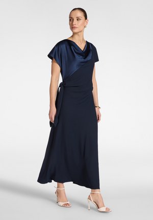 Frau trägt ein marineblaues Wickelkleid mit einem satinierten Wasserfallausschnitt, steht und schaut zur Seite, kombiniert mit weißen Sandalen mit hohen Absätzen.