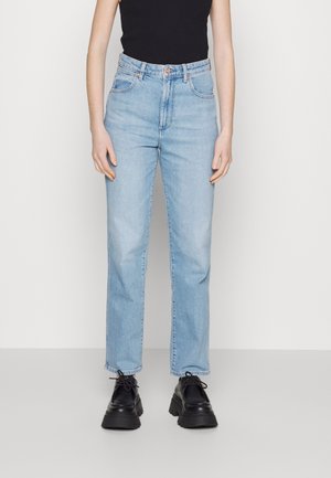 Person iført lyseblå højttaljede straight-leg jeans, sorte chunky snøreløse sko og en sort ærmeløs top mod en ensfarvet baggrund.