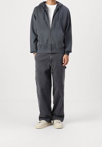 Uomo che indossa una felpa con zip grigio scuro, una t-shirt bianca, pantaloni larghi grigio scuro e sneakers color avorio, in piedi con le mani in tasca.