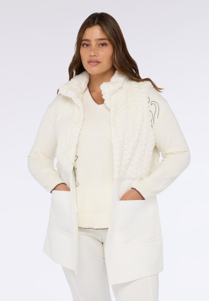 Weißes ärmelloses Oberteil mit einer strukturierten, flauschigen Vorderseite, einfarbigem Rücken und großen Fronttaschen, getragen über einem weißen Strickpullover.