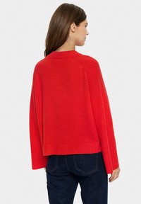 Maglione rosso lavorato a maglia con una trama a coste, maniche ampie e scollo rotondo, abbinato a jeans scuri. La parte posteriore mostra una vestibilità rilassata.