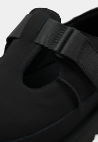 Sandale noire à bout fermé avec une large bride en tissu réglable et un détail ajouré sur la partie supérieure de la chaussure.