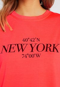 Nainen, joka on pukeutunut korallinväriseen t-paitaan, jossa mustalla tekstillä ovat New Yorkin koordinaatit 40°42'N 74°00'W rintaan painettuna.