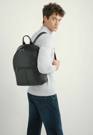 Młody mężczyzna w jasnoszarym swetrze i niebieskich dżinsach, niosący czarny skórzany plecak na jednym ramieniu, spoglądający przez ramię na neutralnym tle.