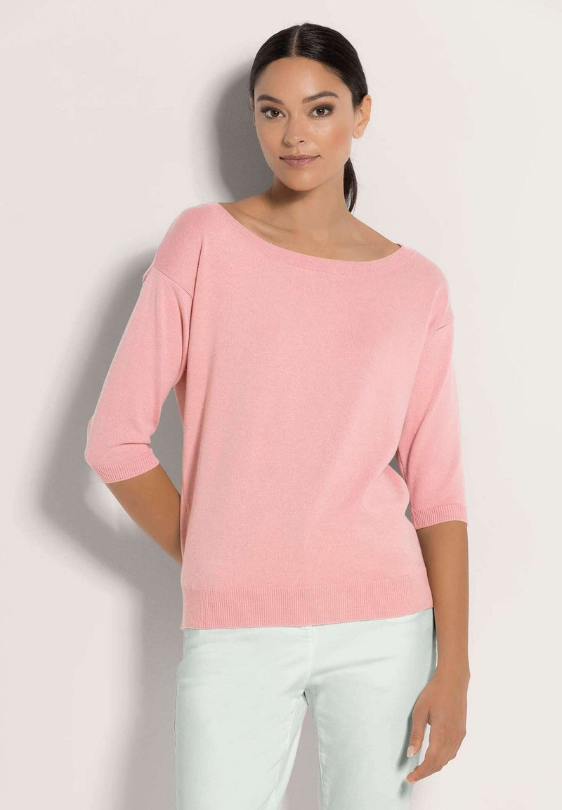 Frau mit dunklem Haar im Pferdeschwanz, die einen hellrosa Boot-Ausschnitt-Pullover mit ellenbogenlangen Ärmeln und hellen Hosen trägt.