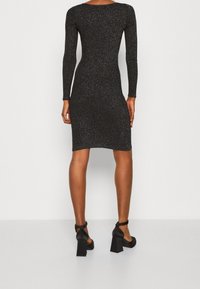 Abito nero attillato a maniche lunghe con texture scintillante, che arriva fino a metà coscia, abbinato a scarpe nere con tacco a blocco.