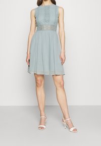 Ljust blå klänning med en strukturerad ovandelsdel som har cirkulära mönster och en genomskinlig kjol. Matchad med vita högklackade sandaler med remmar.