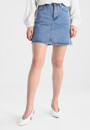 Kvinde iført en blå denim mininederdel med hvide sidelister, en hvid bluse med lange ærmer og hvide slingback sko med hæl.