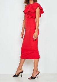 Červené bodycon šaty s jedním ramínkem, zdobené volánkovými akcenty. Hladká látka, délka ke kolenům, v kombinaci s černými vysokými podpatky.