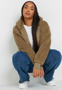 Brun fuskpälsjacka med dragkedja och beige innerfoder, ribbade muddärmar och en mysig huva, kombinerad med blå jeans och vita sneakers.