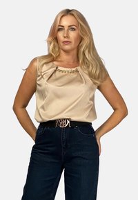 Beżowy top bez rękawów o drapowanym wzorze, z złotym łańcuszkiem przy dekolcie, w zestawieniu z ciemnoniebieskimi spodniami z wysokim stanem i czarnym paskiem.